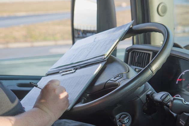 A truck driver sits behind the wheel as they write in a log book.