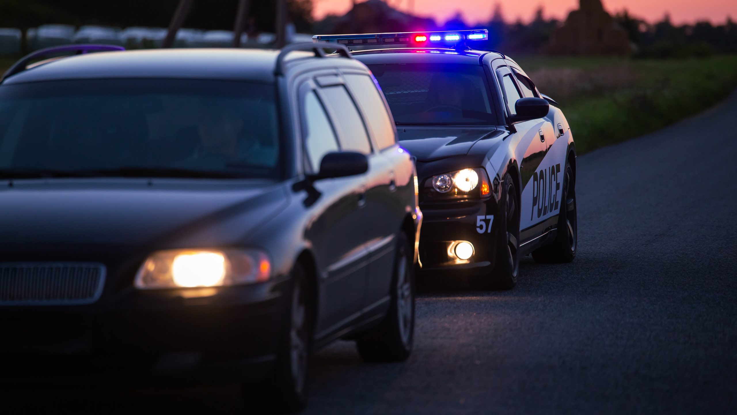 Police car with flashing lights pulling over a sedan at dusk.
