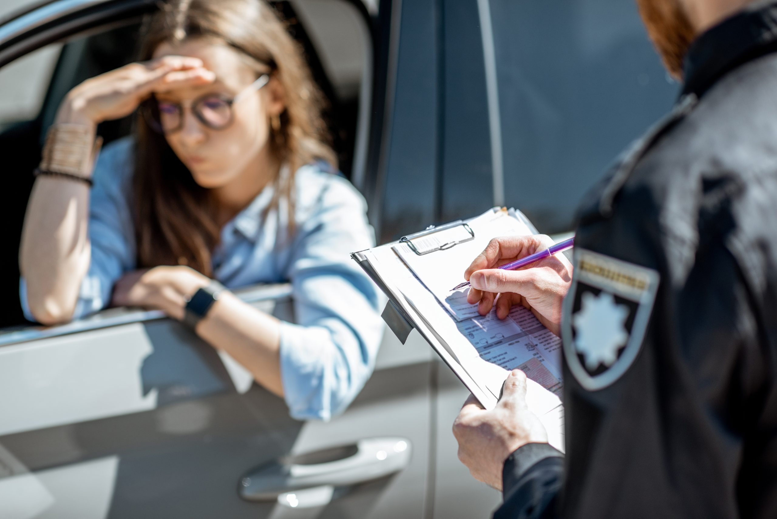 A distressed woman rests her head in her hand at the car window while a police officer writes a ticket on a clipboard.